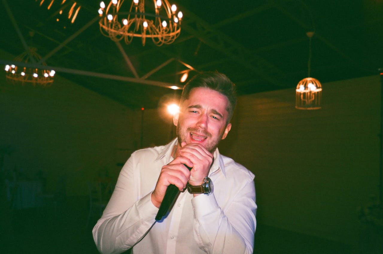 A male singer passionately performing indoors under warm hanging lights, clutching the microphone.